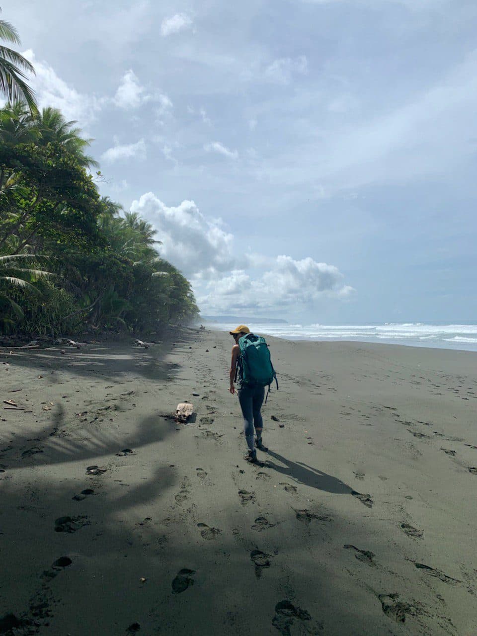 A hiker with a backpack walking a dark volcanic beach lined with palms under a heavy sky, Corcovado National Park